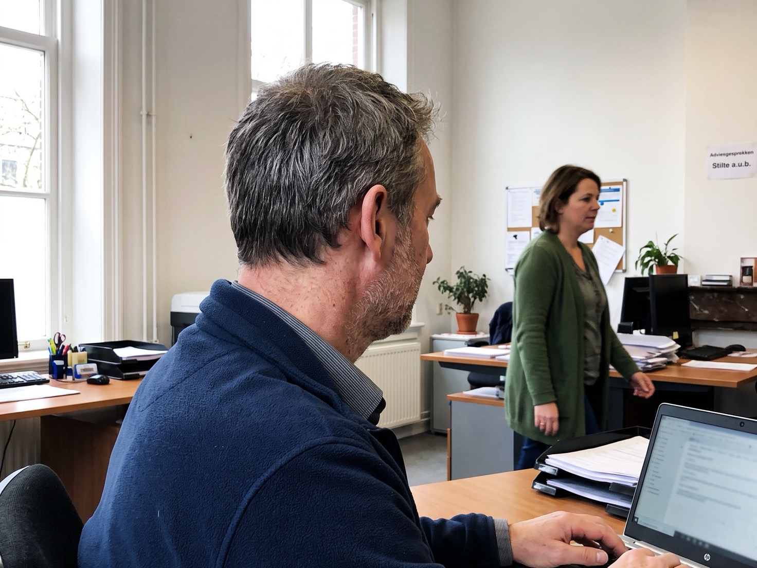Adviseur werkt achter zijn laptop op kantoor, terwijl een collega van de binnendienst op de achtergrond langsloopt
