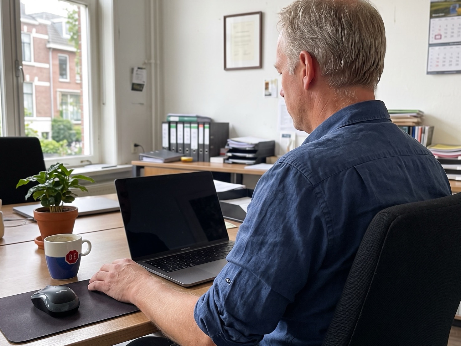 Financieel adviseur van rond de 55 werkt achter zijn laptop in een rustig advieskantoor, van opzij gezien