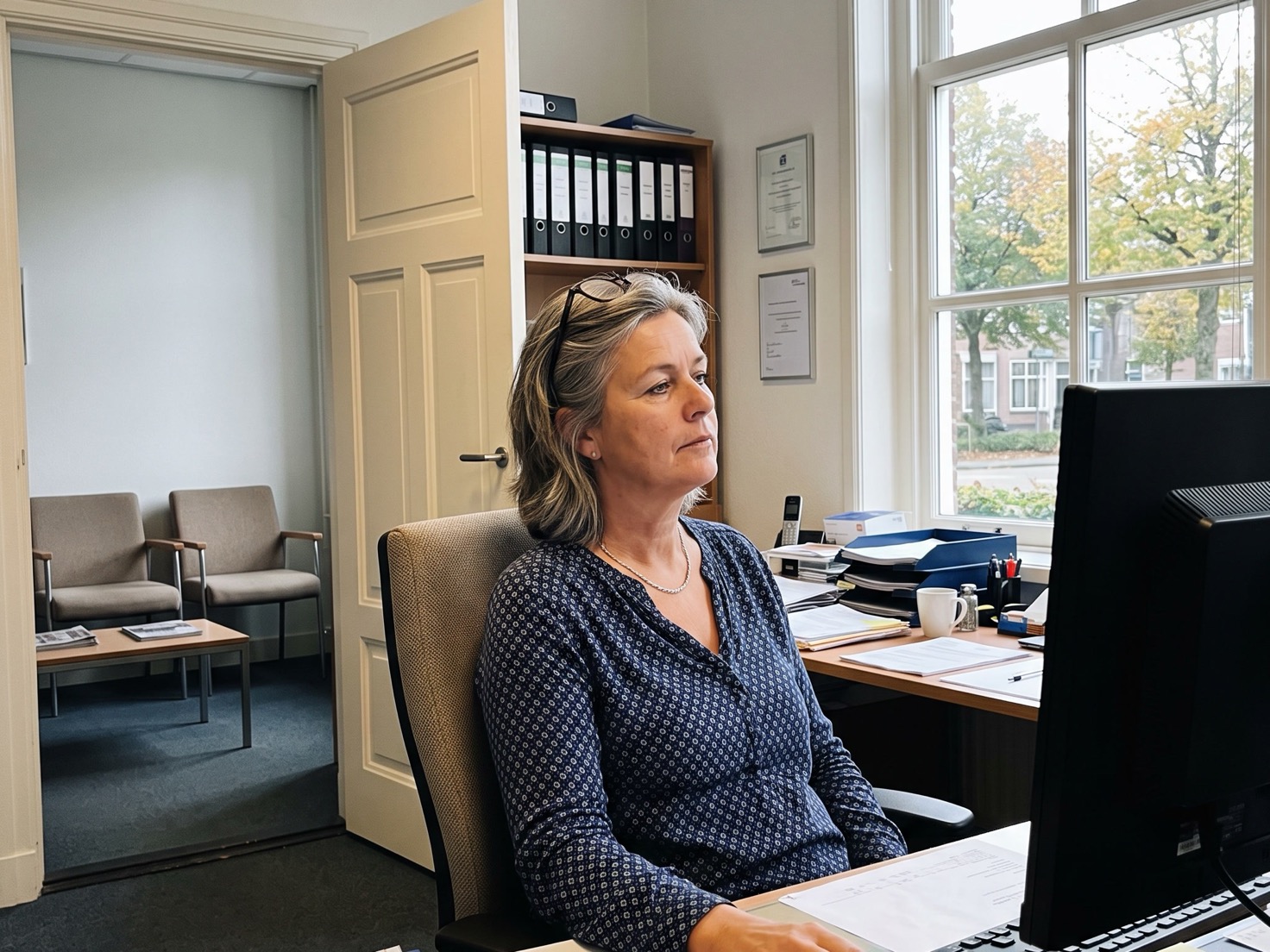 Adviseur werkt aan haar bureau in een rustig advieskantoor met de deur open naar de wachtruimte