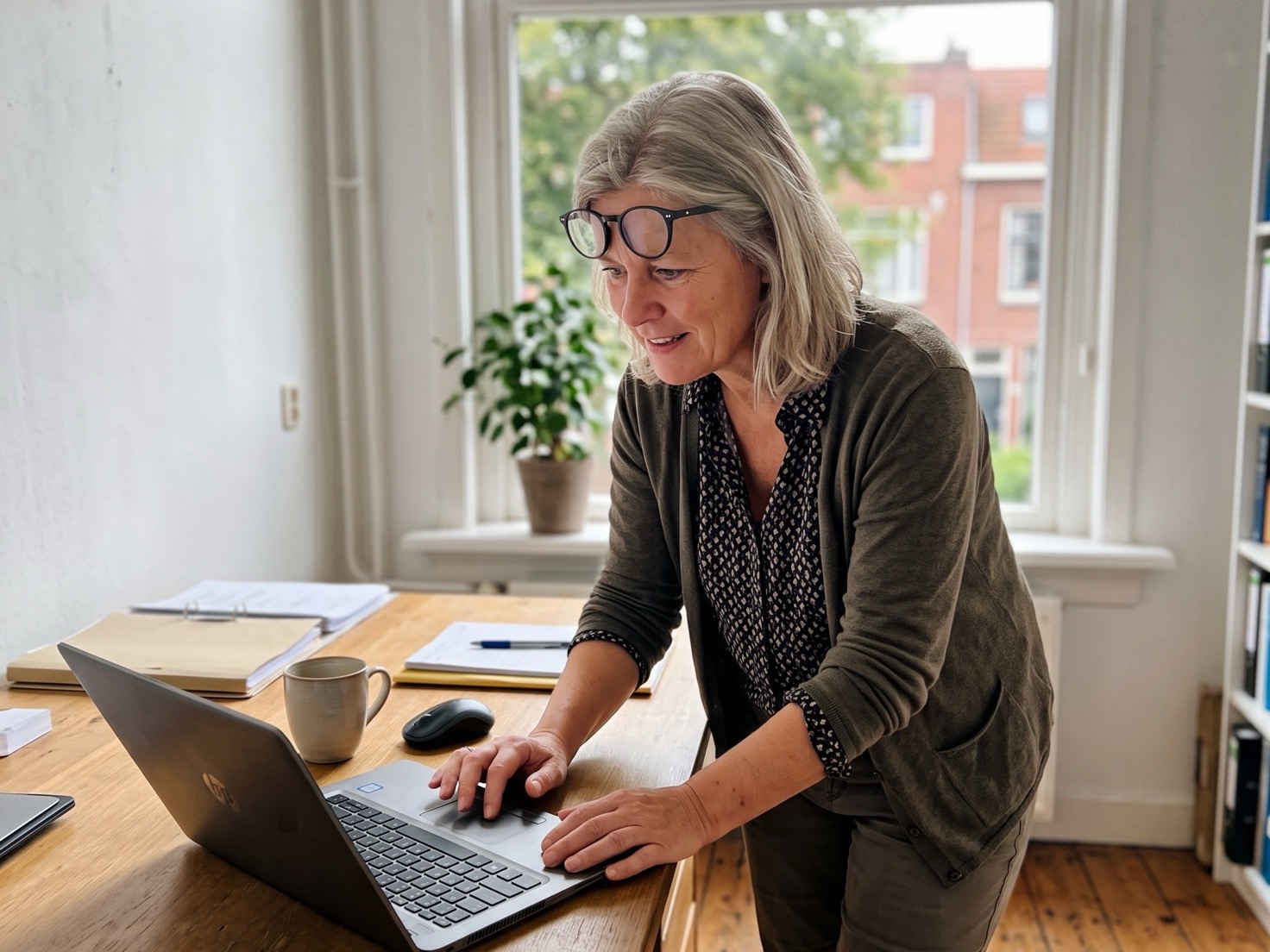 Financieel adviseur van rond de 55 staat naast haar bureau en buigt zich voorover naar haar laptopscherm in een rustig advieskantoor