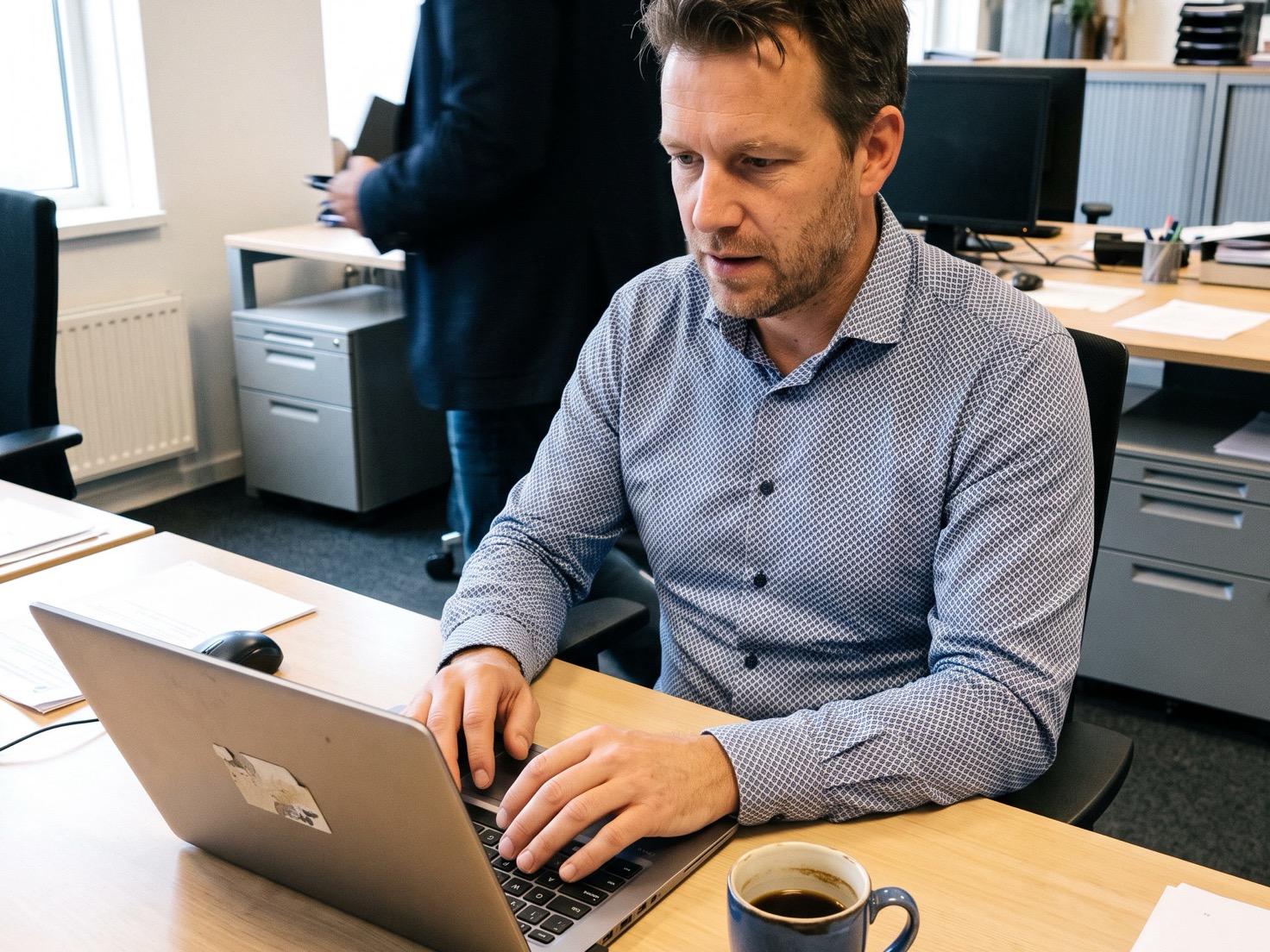 Adviseur typt achter een laptop in een rustig advieskantoor, met een collega op de achtergrond