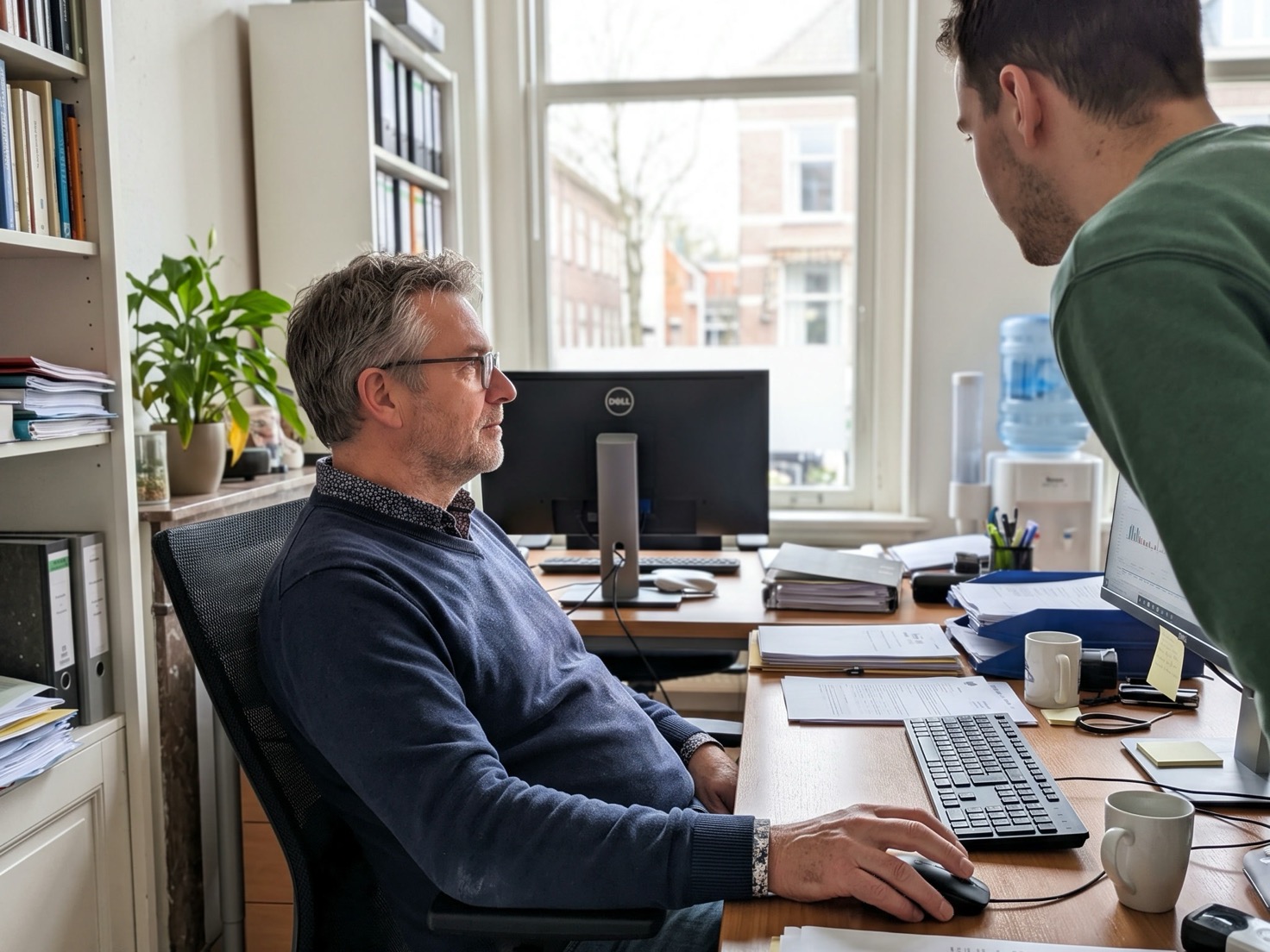 Financieel adviseur werkt achter zijn bureau, een jongere collega kijkt vanaf de achtergrond mee